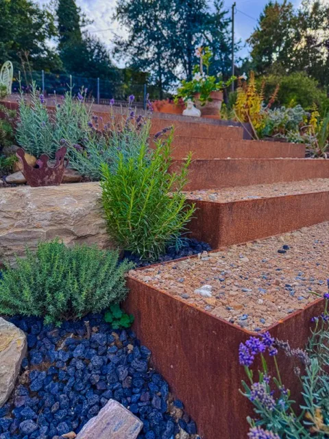 Stufen aus Cortenstahl mit heller Schüttung, dazwischen Lavendel und Rosmarin neben einer Natursteinmauer.